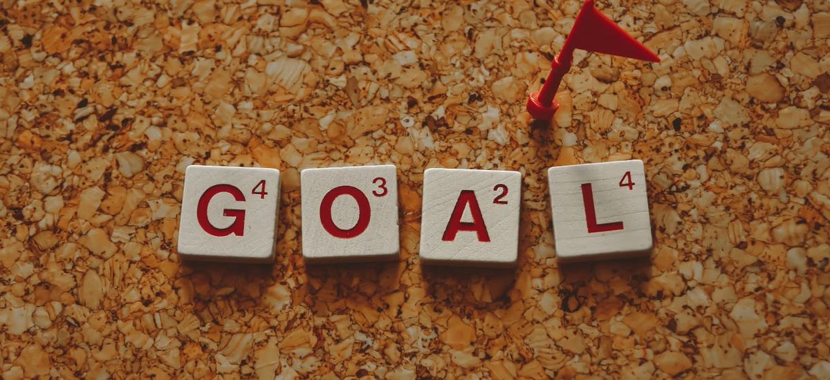 Scrabble tiles forming the word 'goal' with a small red flag on a cork board background.