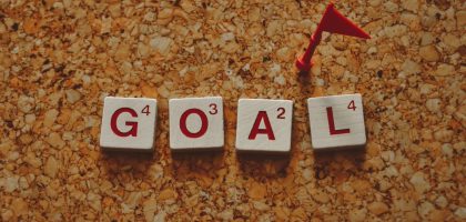 Scrabble tiles forming the word 'goal' with a small red flag on a cork board background.