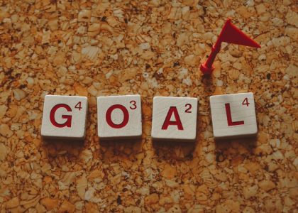 Scrabble tiles forming the word 'goal' with a small red flag on a cork board background.